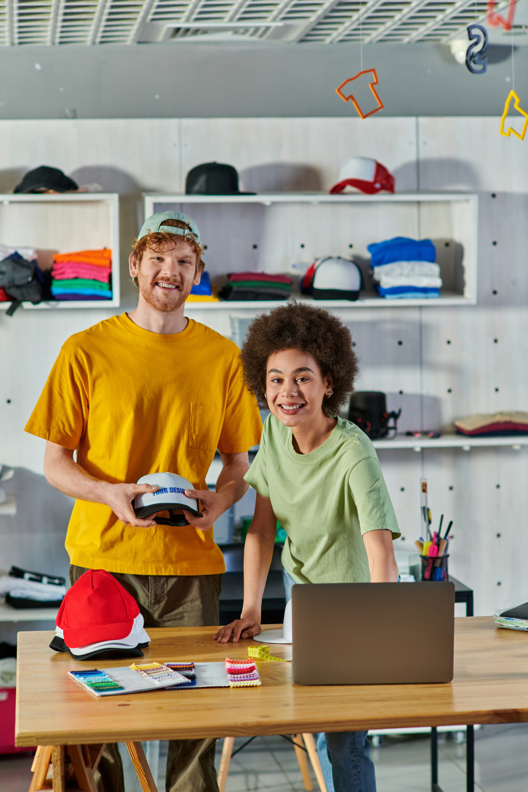 Cheerful young multiethnic designers looking at camera while working with snapbacks, laptop and cloth samples on table in print studio at background, sustainable business model concept